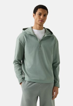 Ung mand iført en lysegrøn hættetrøje med halv lynlås og matchende bukser, stående med den ene hånd i lommen.