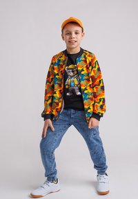 Chłopiec w pomarańczowej czapce, kolorowej kurtce w kamuflażu, czarnej koszulce z grafikiem niedźwiedzia, niebieskich dżinsach i białych trampkach stoi z nogami rozstawionymi.