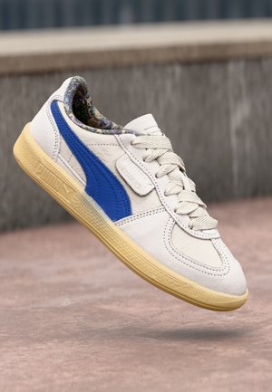 White leather sneaker with blue side stripe, textured accents, and light yellow rubber sole. Features patterned inner lining and standard laces.