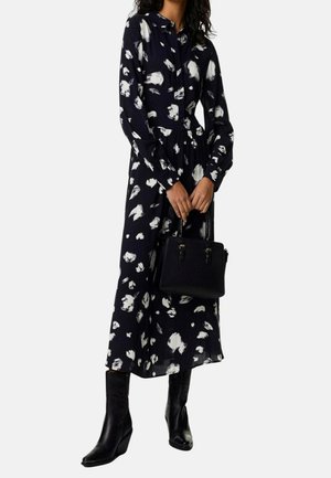 Frau trägt ein langärmeliges Kleid mit schwarzem und weißem Muster, schwarze Stiefel und hält eine schwarze Handtasche vor einem einfarbigen Hintergrund.