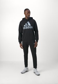 Sudadera negra con gran logotipo azul de Adidas, combinada con pantalones de chándal negros y zapatillas atléticas grises. Tejido suave, corte relajado, puños elásticos.