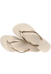 Beige flip-flops dengan sol bertekstur dan tali berwarna emas yang mengkilap dengan pola yang halus. Desainnya ringan dan cocok untuk dikenakan santai.