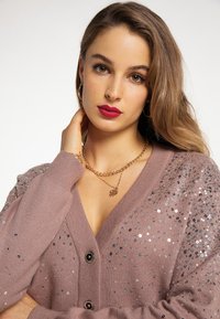 Tânără cu păr lung, castaniu deschis, îmbrăcată cu un cardigan mauve cu paiete, cercei aurii rotunzi, lanțuri de aur suprapuse și ruj roșu.