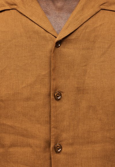Κοντινό πλάνο καφέ λινου πουκαμίσου με τρία ορατά κουμπιά και μερικώς ανοιχτό γιακά που αποκαλύπτει τριχοφυΐα στο στήθος.
