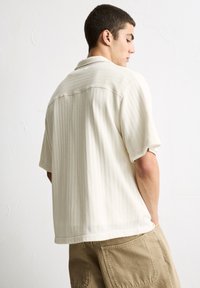 Short-sleeve, collared shirt in cream, textured fabric with a subtle pattern. Casual fit, featuring a straight hem and no visible hardware.