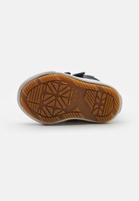 Zapatilla negra con suela de goma, que presenta una suela exterior de color goma texturizado, entresuela blanca y tiras de Velcro para el cierre.