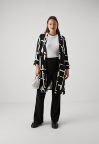 Fekete-fehér mintás kimono széles ujjal, fehér felsővel és fekete, bő szárú nadrággal párosítva. A modell egy kis ezüst táskát tart.