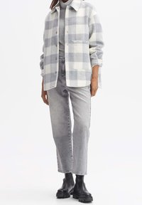 Chaqueta de forro polar a cuadros gris y blanco con cierre de botones, bolsillo frontal, llevada sobre un suéter de cuello alto gris y combinada con unos jeans grises y botas negras.