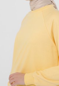 Sudadera amarilla con tejido suave, mangas raglán y puños de canalé. La camiseta tiene un corte holgado y una textura suave, sin patrones visibles.