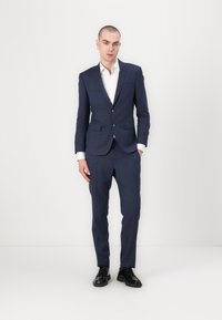 Navy blue suit with a notched lapel, two buttons, and slim fit design. White shirt under the blazer, paired with black dress shoes.