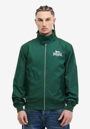 Young man with braided hair wearing a dark green zipped jacket with "Lonsdale London" logo and blue jeans, standing against a white background.
