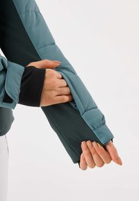 Turquoise donzen jas met een gestroomlijnd ontwerp, met een contrasterende zwarte mouw en een hand die de manchet vasthoudt. De stof lijkt gevuld en glad.