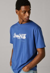 Μπλε βαμβακερό μπλουζάκι με κοντά μανίκια, στρογγυλή λαιμόκοψη και λευκό λογότυπο τυπωμένο. Διαθέτει άνετη εφαρμογή και μαλακή υφή.