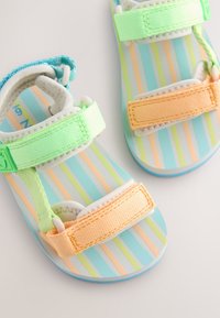 Sandali per bambini con cinturini in azzurro chiaro, con chiusure regolabili in Velcro verde e arancione e un motivo a strisce colorate sulla soletta.
