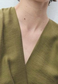 Tissu texturé vert olive avec un col en V. L'encolure révèle une peau lisse, avec de petits grains de beauté visibles sur le cou.