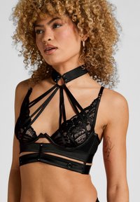 Μαύρο εσώρουχο με δαντελένια κύπελλα, σατέν λουριά και κολάρο με μεταλλικές λεπτομέρειες. Ο σχεδιασμός περιλαμβάνει διασταυρούμενες λεπτομέρειες και ρυθμιζόμενα στοιχεία.