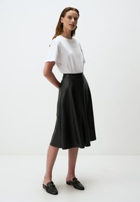 Jupe midi en cuir noir avec un design évasé, taille haute et texture lisse, associée à un t-shirt blanc et des mocassins noirs.