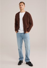 Cárdigan marrón de punto con cierre de botones sobre una camiseta blanca, combinado con jeans de color azul claro y zapatos negros. Fondo liso.