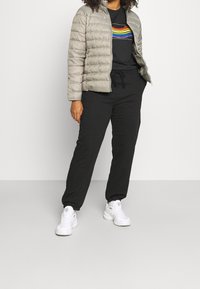 Hellgraue Pufferjacke, schwarzes Grafik-T-Shirt mit Regenbogenstreifen, schwarze Jogginghose und weiße Sneakers, stehend vor einem schlichten Hintergrund.