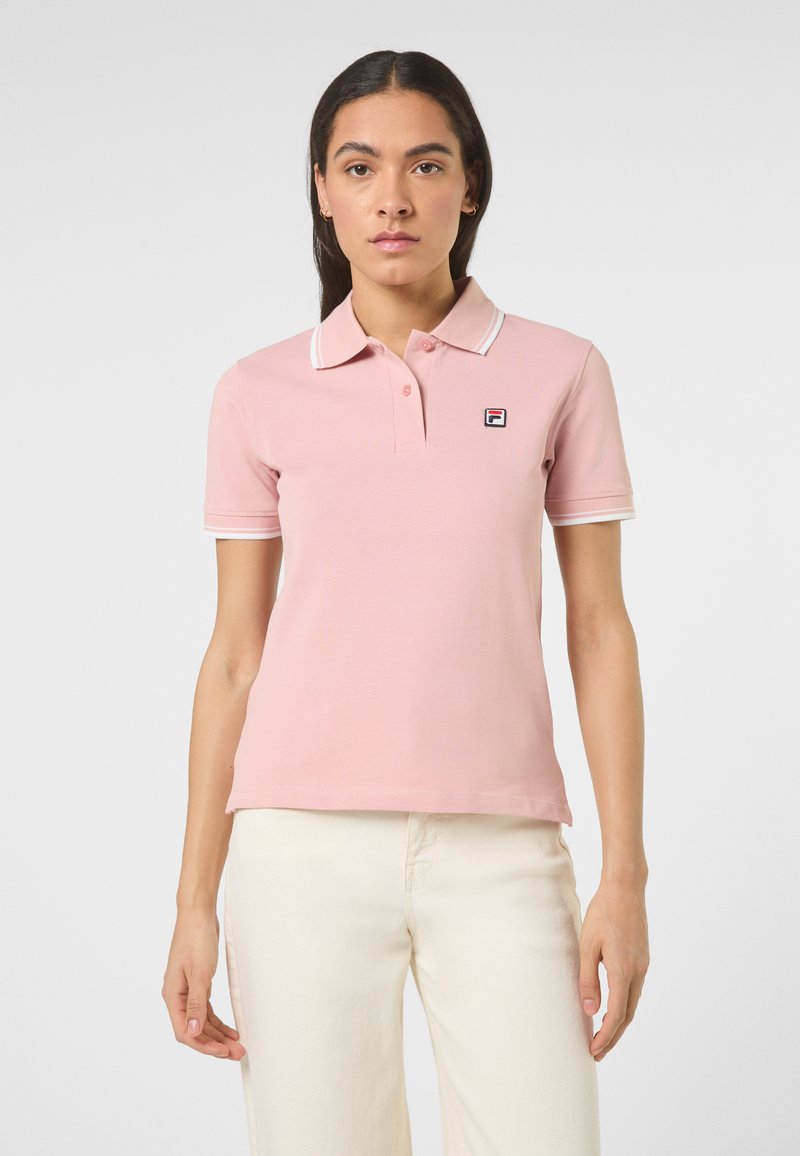 Kvinde iført lyserød kortærmet poloshirt med logo på brystet og hvide bukser, stående mod en ensfarvet lys baggrund.