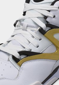 Zbliżenie białego trampka z białymi sznurowadłami oraz czarno-złotymi plastikowymi nakładkami na skórzanej i siateczkowej cholewce.