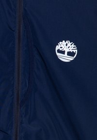 Navy blue jacket features a front zipper, a side pocket, and a white tree logo on the chest, made from a smooth, lightweight fabric.