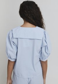 Blusa azzurro chiaro con texture, caratterizzata da un ampio colletto e maniche a sbuffo, con un taglio morbido e un design semplice. Finitura in tessuto liscia.