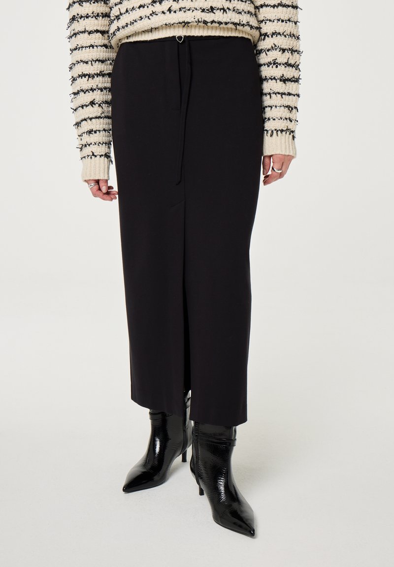 Zwarte, middellange rok van soepel materiaal, met een voorkruising en een verstelbare strik. Draag het met glanzende zwarte enkelboots.