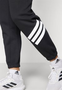 Pantaloni della tuta neri con caviglie affusolate presentano tre strisce bianche sulla gamba sinistra. Indossati con scarpe bianche e calze nere con ricamo adidas.