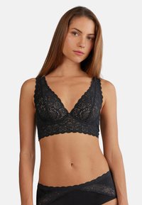 Zwart kanten bralette met driehoekige cups, gegolfde randen en brede banden. Heeft ingewikkelde bloemenpatronen en een zachte, gestructureerde afwerking.