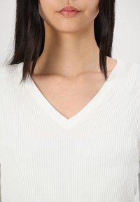 White ribbed v-neck sweater with a textured surface. The neckline is subtly defined, leading to a fitted look around the upper body.
