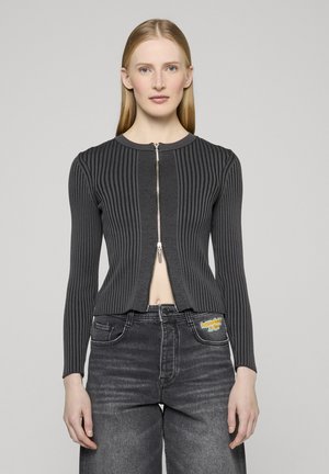 Donna con capelli biondi lisci che indossa un maglione grigio scuro a costine con zip e jeans neri sbiaditi con una toppa sulla tasca.