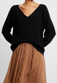 Osoba v černém pleteném svetr s V-výstřihem a světle hnědé sukně s knoflíky a malým černým tečkovaným vzorem, stojící na jednobarevném pozadí.