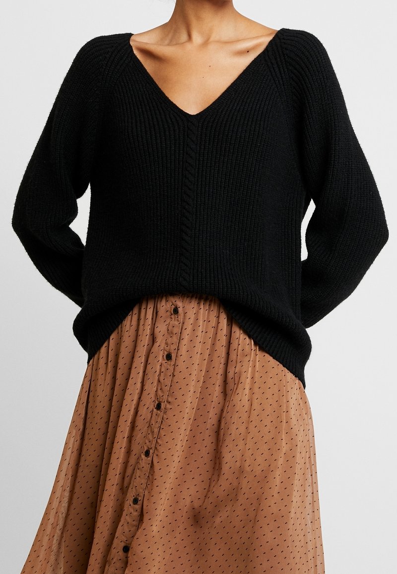 Osoba v černém pleteném svetr s V-výstřihem a světle hnědé sukně s knoflíky a malým černým tečkovaným vzorem, stojící na jednobarevném pozadí.