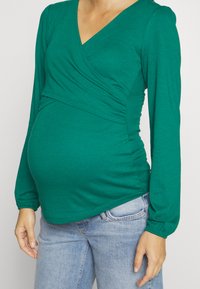 Zöld, hosszú ujjú kismama felső puha anyagból, keresztbe álló V-nyakkal és ráncolt részletekkel a deréknál, kék farmernadrággal párosítva.