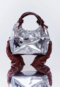 Mains gantées de cuir rouge tenant un petit sac à main argenté brillant avec le haut froncé et des anses tressées rouge foncé au-dessus d'une surface réfléchissante.