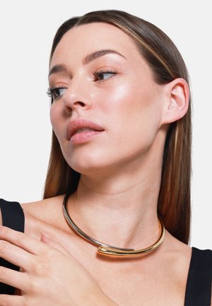 Gold- und Silbermetall-Choker-Halskette mit einem glatten, geschwungenen Design und einer spitzen Akzentuierung. Das Model trägt sie über einem schwarzen Oberteil.