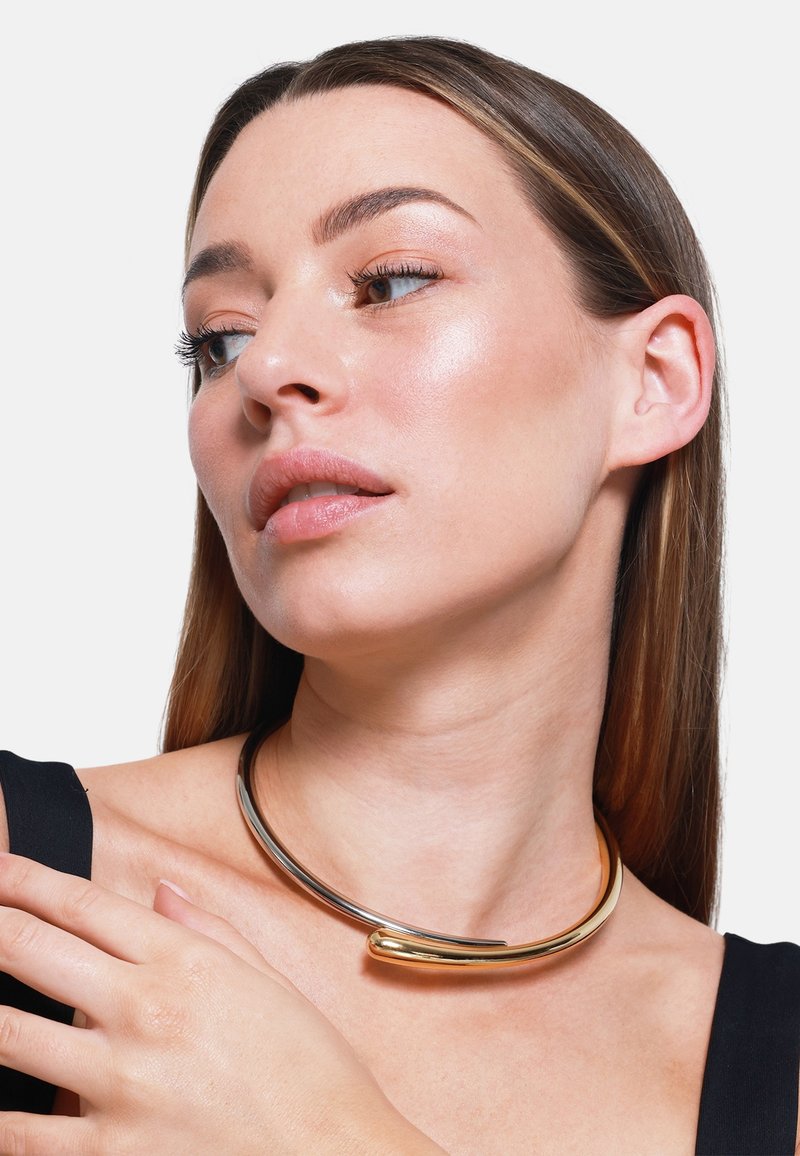 Guld- og sølvmetal choker halskæde med et glat, buet design og en spids accent. Modellen bærer den over en sort top.