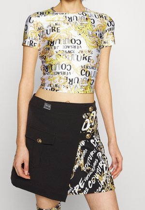 Jonge vrouw die een witte en gouden cropped T-shirt met een sierlijk patroon draagt, en een zwarte minirok met een bijpassend paneel met print en gouden knopen.