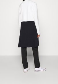 Persona che indossa un lungo cardigan color block bianco e nero, pantaloni neri e sneakers bianche Converse, in piedi di fronte a uno sfondo neutro.