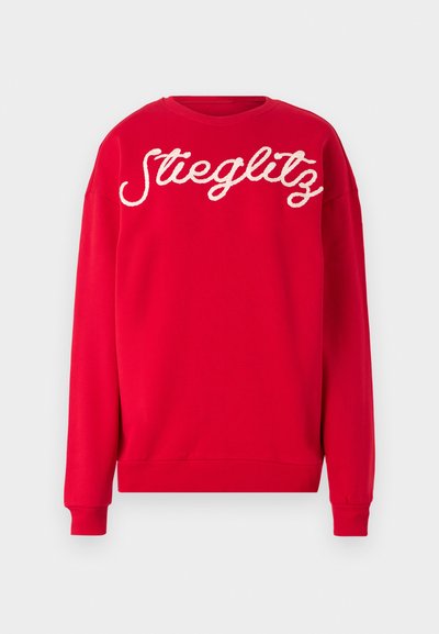 Rød hettegenser med hvit, fluffy brodert tekst "Stieglitz" på forsiden. Lange ermer og ribbestrikkede mansjetter. Myk stofftekstur.