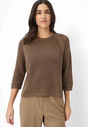 Kvinde med mørkt hår iført en brun, tekstureret striktrøje med trekvartlange ærmer og beige bukser, stående mod en hvid baggrund.