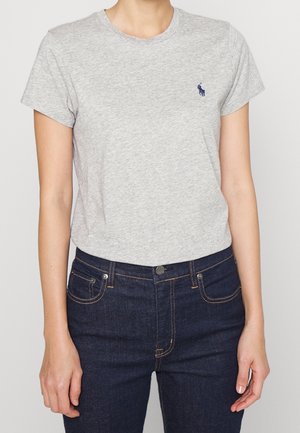 T-shirt grigia in cotone con maniche corte e scollo rotondo, con un piccolo logo blu navy sul petto a sinistra, abbinata a jeans in denim scuro.