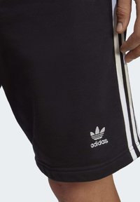 Juodi sportiniai šortai, pagaminti iš minkštos medžiagos, su trimis baltais vertikaliais dryžiais šoninėse pusėse ir mažuoju balto siuvinėjimo logotipu.