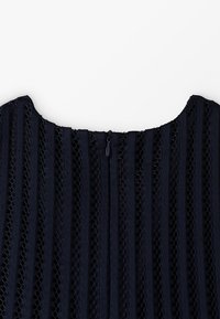 Tummansininen mekko, jossa on pystysuorat raidat ja läpikuultavia verkko-osia. Pyöreä pääntie ja takana vetoketjukiinnitys.