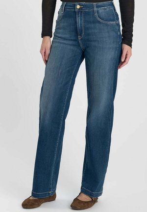 Frau trägt blaue Jeans mit geradem Bein, schwarzen langärmligen Oberteil und braune flache Schuhe und steht vor einem einfarbigen Hintergrund.
