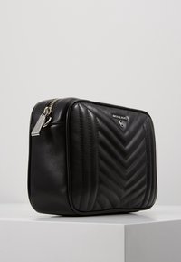 Schwarze, gesteppten Lederhandtasche mit Chevron-Muster, silberfarbener Hardware-Reißverschluss und Logo-Detail auf der Vorderseite. Kompakte rechteckige Form.