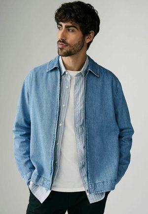 Ljusblå denimjacka med en avslappnad passform. Under jackan bärs en randig skjorta med knappar i mjuk blå och vit. Enkel, modern design.