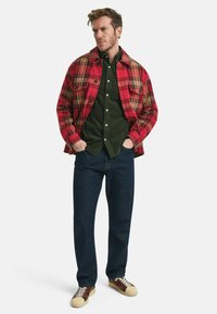 Camisa de franela a cuadros roja y amarilla sobre una camisa de botón verde oscuro, combinada con jeans de mezclilla oscura y zapatillas marrones. Dos bolsillos en el pecho de la camisa de franela.