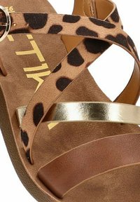 Sandalia con estampado de leopardo, parte superior de ante marrón, tira metálica dorada y suela plana. Tiras cruzadas para un ajuste seguro; materiales texturizados.